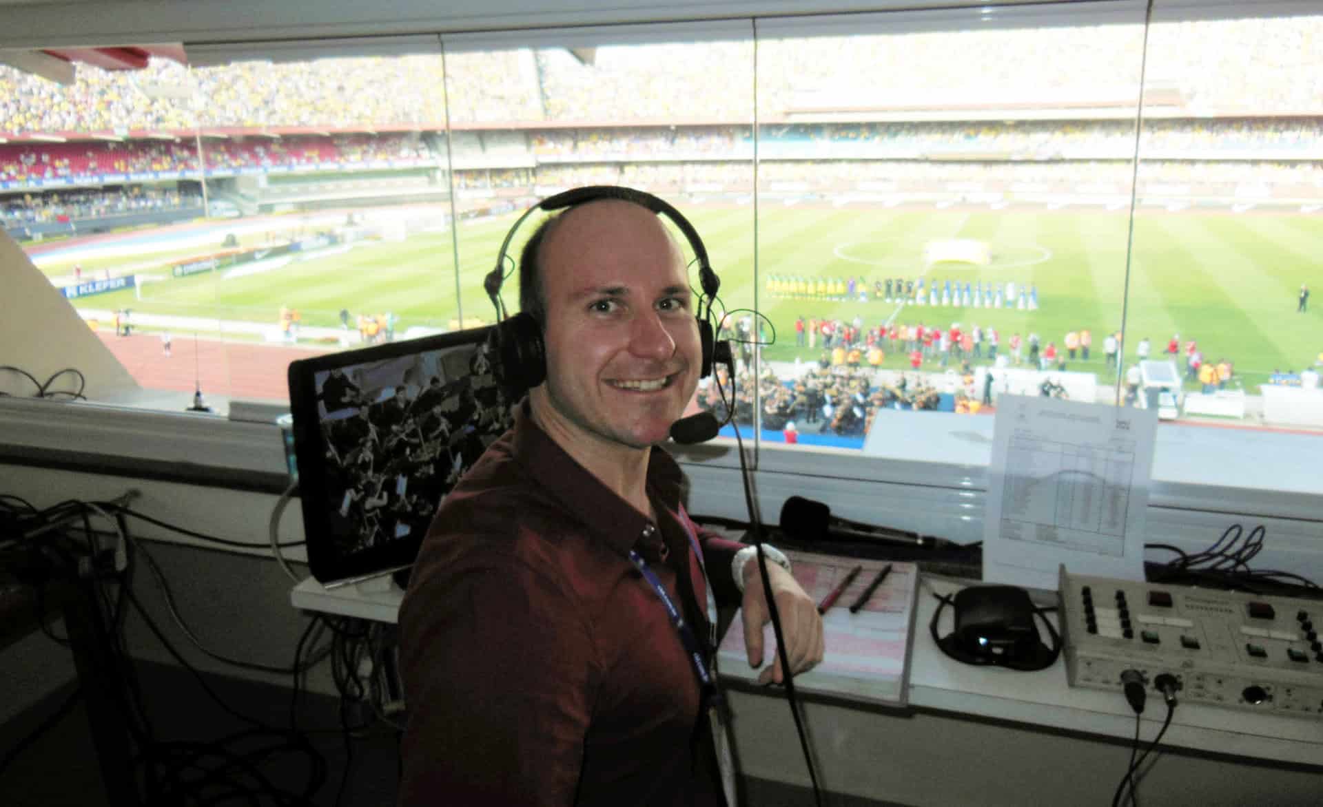 Broadcast sports commentator Duane Dell'Oca in a press box overlooking a vibrant stadium with a football field during a live event.
