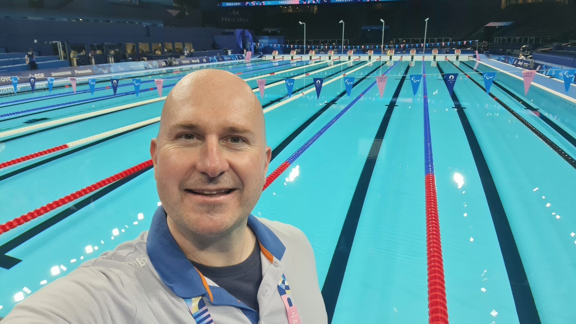 Competitive swimmer at the Paris 2024 Paralympic swimming venue, smiling with Olympic-sized pool lanes in the background.