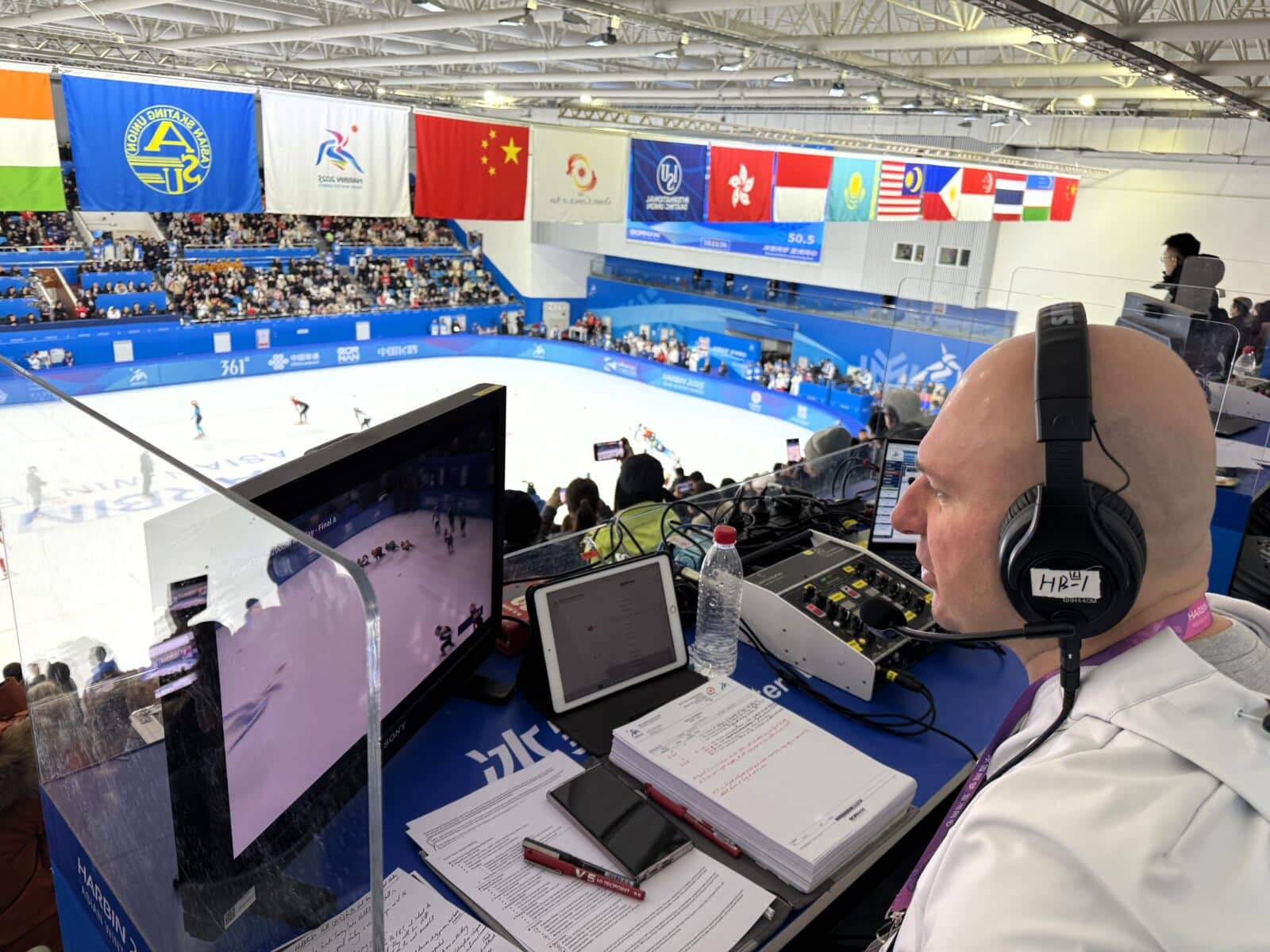 Broadcasting ice skating event at the Winter Olympics with professional equipment and commentary setup, showcasing the vibrant atmosphere and international flags in the background.